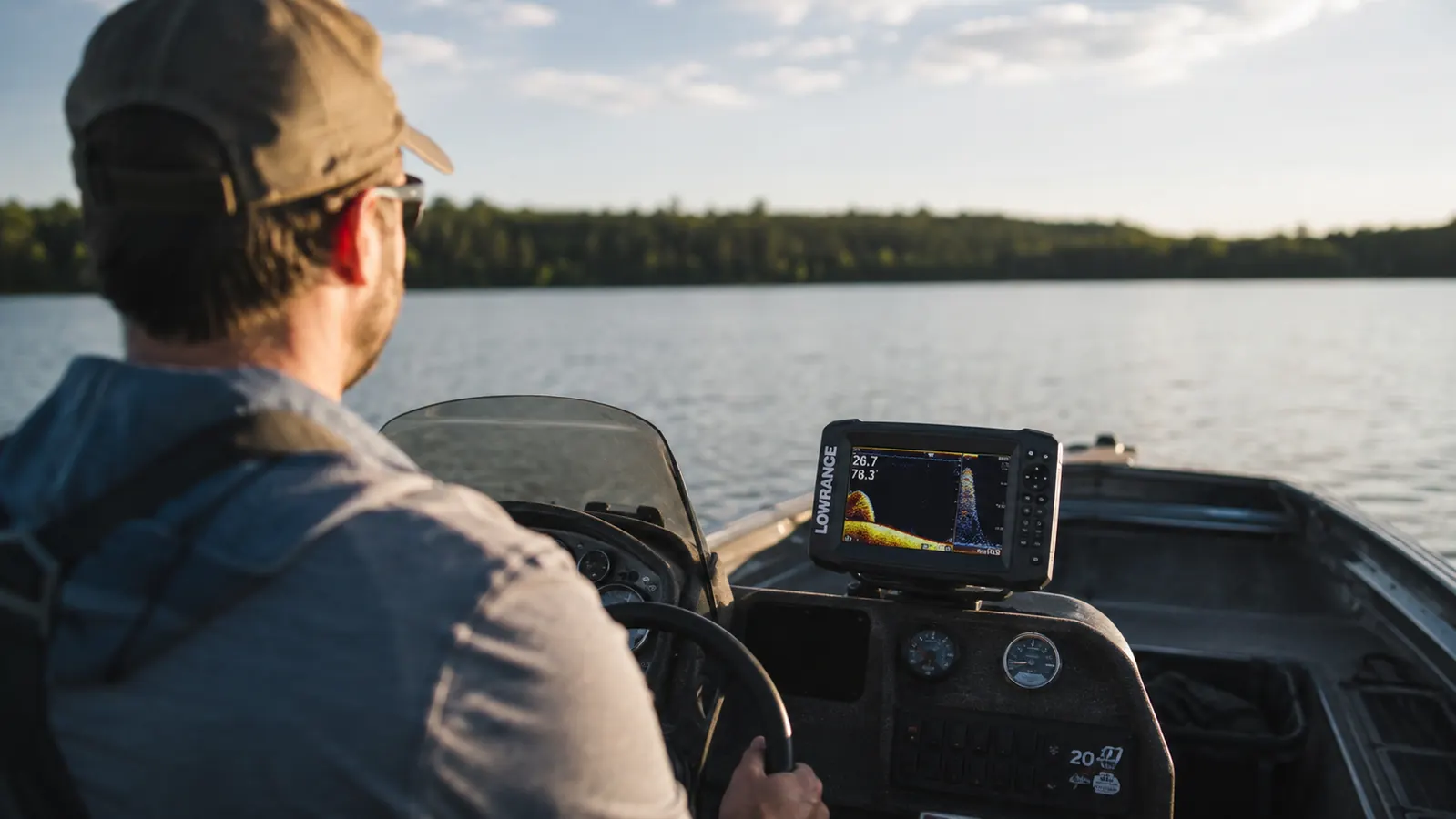 A small fish finder mounted at the console of an aluminum boat, seen over the driver's shoulder on a calm lake in late afternoon light
